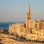 View of Valletta’s historic limestone buildings and church spire overlooking the Mediterranean Sea at sunset, a scene travelers enjoy when heading to Valletta from Malta Airport.