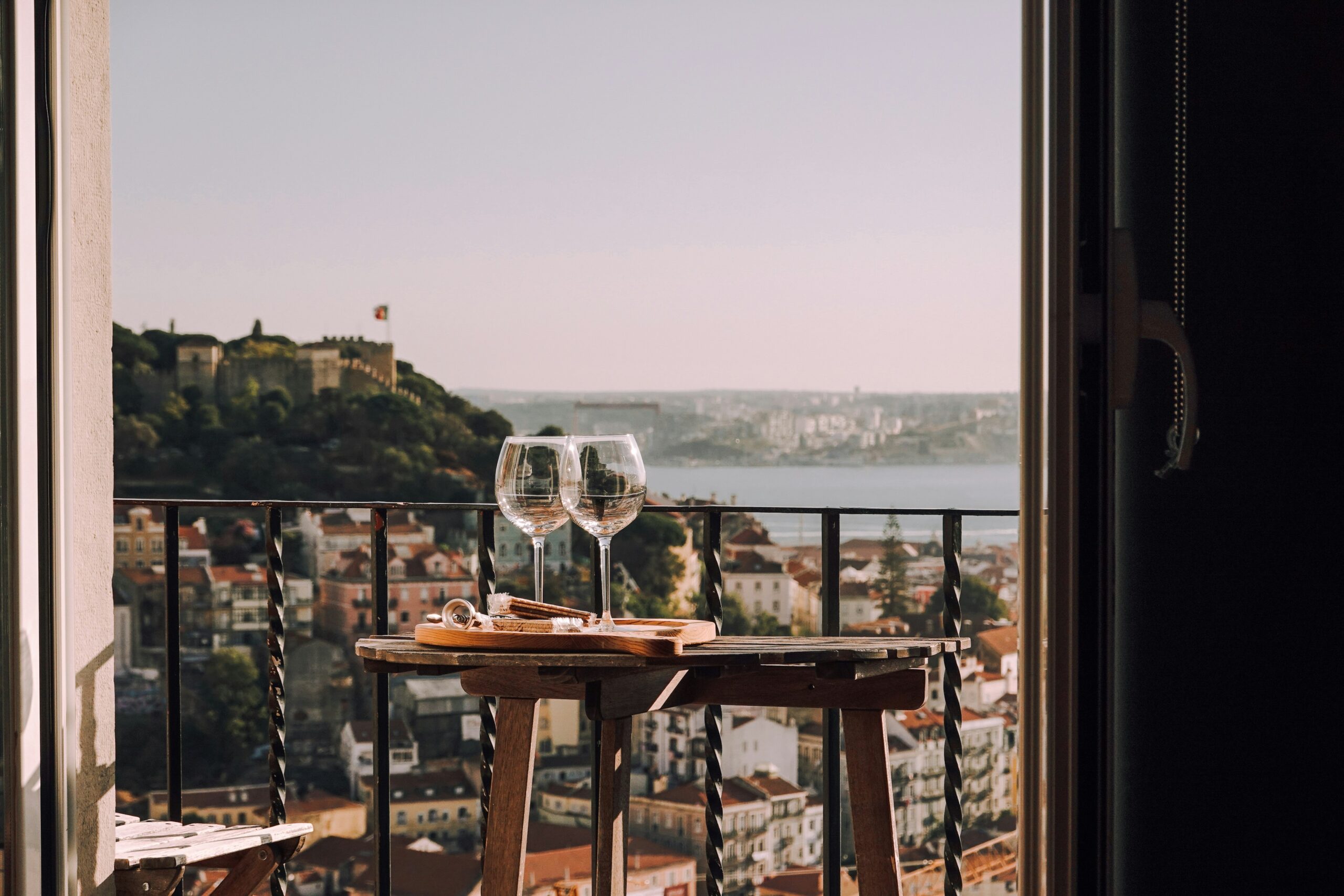 two wine glasses atop a table overlooking a balcony and Lisbon's neighborhoods, water, and hills