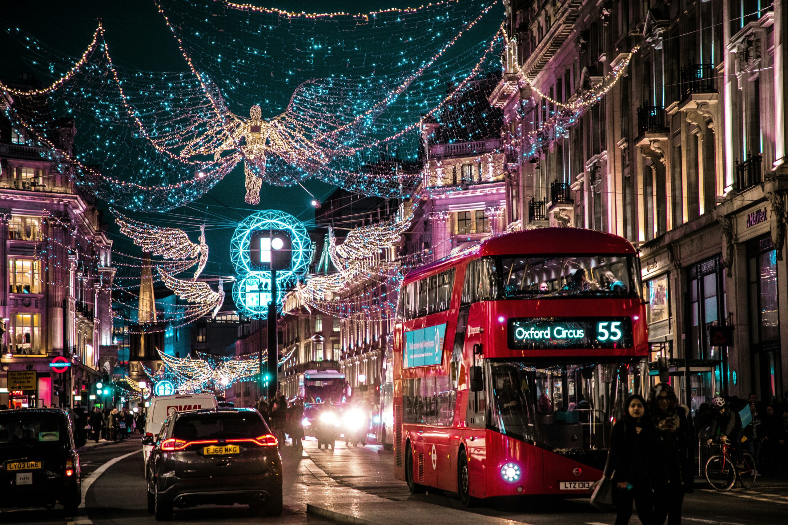 A busy street lit up with Christmas lights shaped like angels with a big red bus and lots of cars.