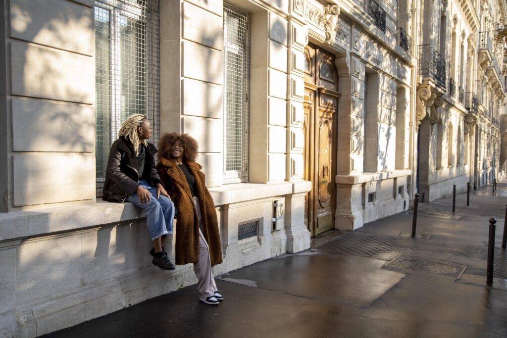 Two women in stylish winter coats talking outdoors on a sunny European city street.