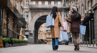women enjoying a shopping trip in Milan, dressed in elegant coats and carrying colorful shopping bags along a cobblestone street.