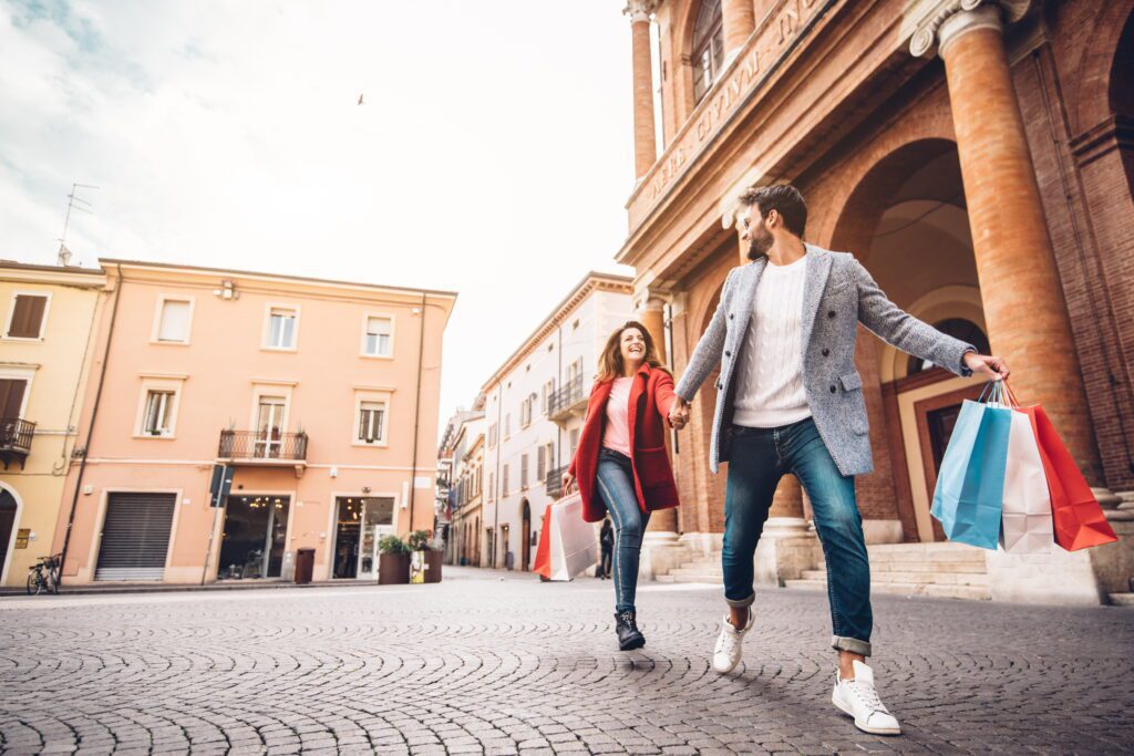 A happy couple holding hands and carrying shopping bags while running through a sunny Italian plaza.