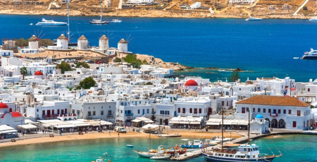 Panoramic aerial view of Mykonos Town (Chora), showcasing the dense cluster of whitewashed Cycladic buildings with red-domed churches, a busy waterfront harbor with traditional wooden boats moored in turquoise water, and the iconic row of five windmills standing on the hilltop overlooking the deep blue Aegean Sea, with luxury yachts anchored in the distance.