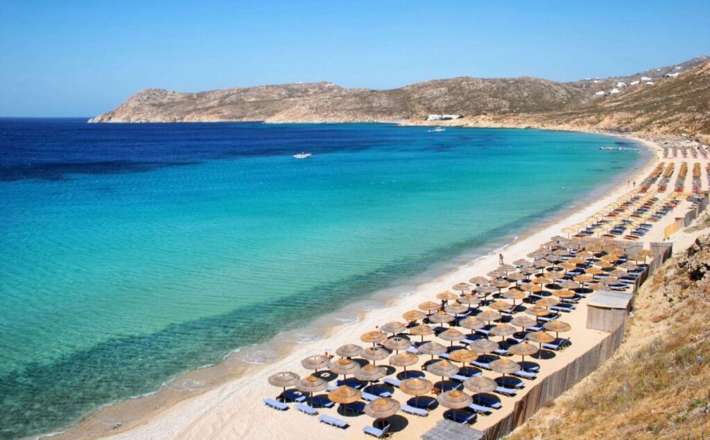 Aerial view of a long, curved sandy beach in Mykonos on a clear summer day, with neatly arranged rows of thatched straw umbrellas and sun loungers stretching along the shore, vivid turquoise and deep blue Aegean water forming a striking color gradient, and a rugged, arid hillside headland closing off the bay in the background.