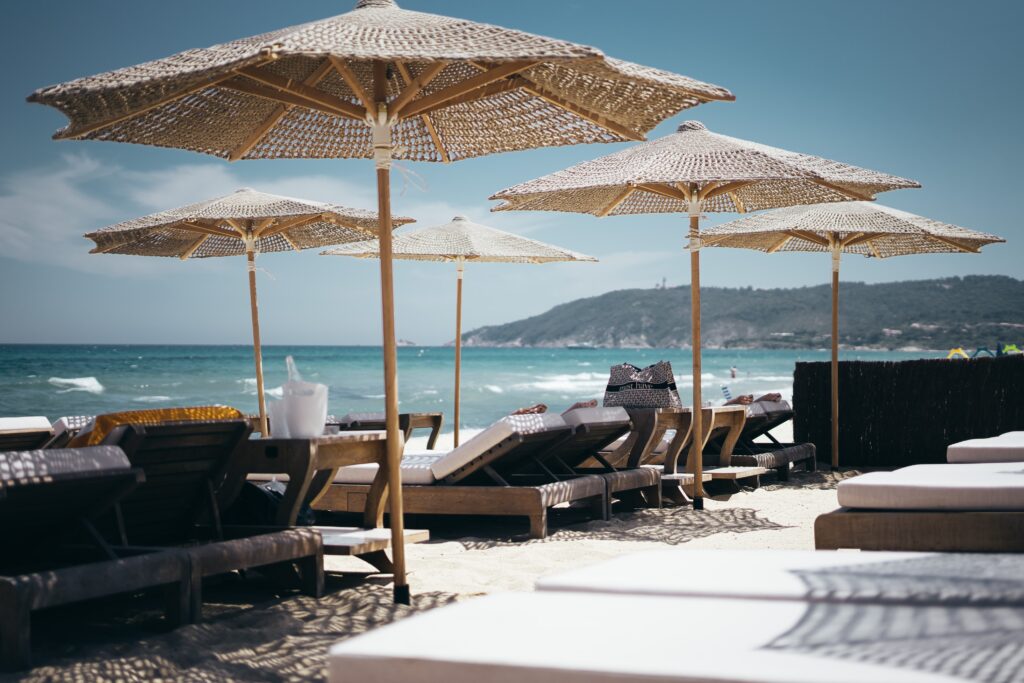 Close-up view of a luxury beach club setup in Mykonos, featuring cushioned wooden sun loungers arranged under large woven rattan umbrellas casting intricate shadows on the sand, with a choppy turquoise Aegean Sea and a green hilly headland visible in the background under a bright summer sky.