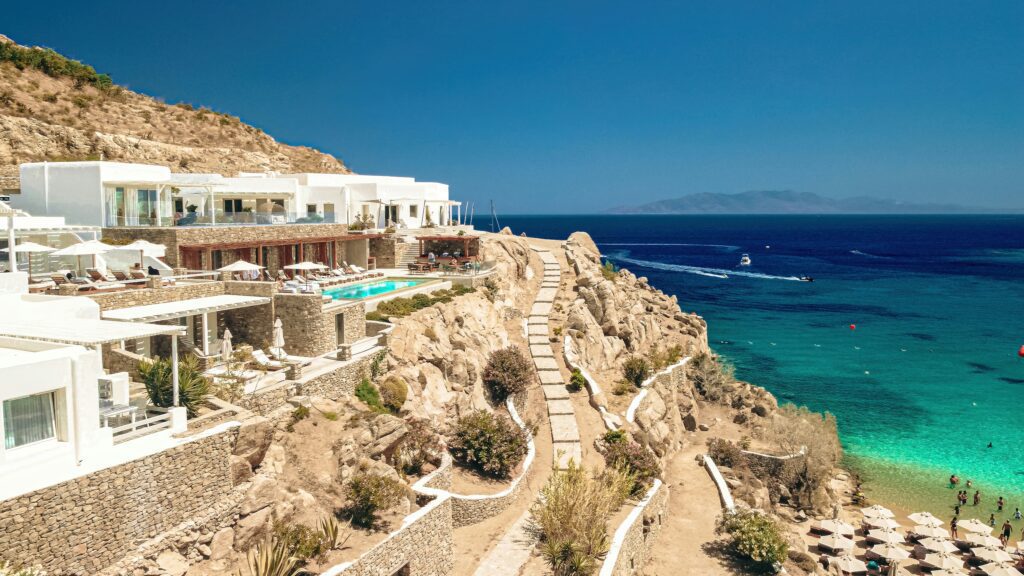 Aerial view of a cliffside luxury hotel in Mykonos, with whitewashed multi-level architecture and stone terraces carved into the rocky hillside, featuring an infinity pool, sun loungers with umbrellas, and stone stairways leading down to a small sandy cove where swimmers enjoy the crystal-clear turquoise water, with a deep blue Aegean Sea and a distant island silhouette on the horizon.