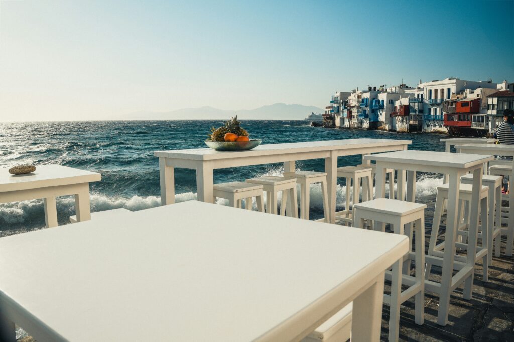 Street-level view of an outdoor waterfront bar in Little Venice, Mykonos, with white high-top tables and stools set directly beside the crashing waves of the choppy Aegean Sea, a bowl of fresh tropical fruit centered on one table, and the colorful neoclassical houses of Little Venice with blue and red balconies visible in the background under a hazy midday sky.