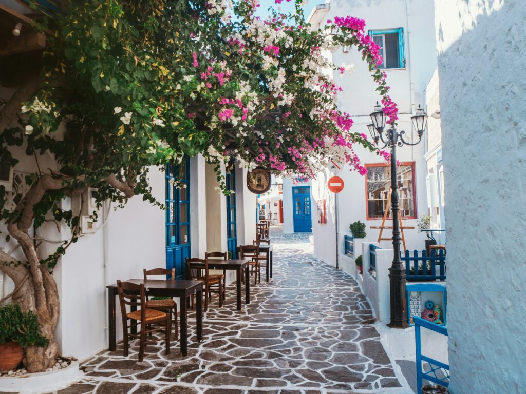 Charming narrow cobblestone alley in Mykonos Town, lined with whitewashed buildings featuring bright blue doors and window frames, a blooming bougainvillea tree with vivid pink and white flowers arching over an outdoor taverna with wooden tables and chairs, a vintage iron street lamp, and small shops receding into the sun-dappled pedestrian lane in the background.