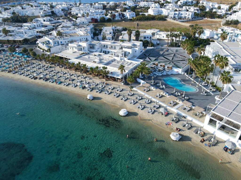 Aerial view of a luxury beachfront hotel in Mykonos, featuring rows of sun loungers and umbrellas lining a sandy beach with crystal-clear turquoise water, an outdoor oval swimming pool surrounded by palm trees, and the iconic whitewashed Cycladic architecture of the town stretching across the hillside in the background.
