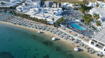 Aerial view of a luxury beachfront hotel in Mykonos, featuring rows of sun loungers and umbrellas lining a sandy beach with crystal-clear turquoise water, an outdoor oval swimming pool surrounded by palm trees, and the iconic whitewashed Cycladic architecture of the town stretching across the hillside in the background.