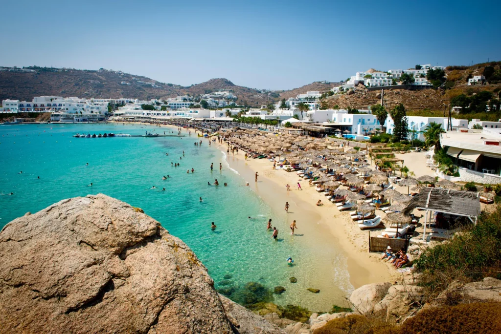 Elevated view of a busy sandy beach in Mykonos on a sunny summer day, with swimmers enjoying the shallow crystal-clear turquoise water, rows of sun loungers shaded by thatched straw umbrellas lining the shore, and whitewashed Cycladic hotels and villas scattered across the dry hillside in the background, shot from a rocky foreground vantage point.