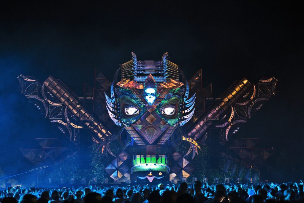 The impressive robotic tribal face stage at Tomorrowland festival with glowing white eyes and a skull symbol, overlooking a large crowd at night.