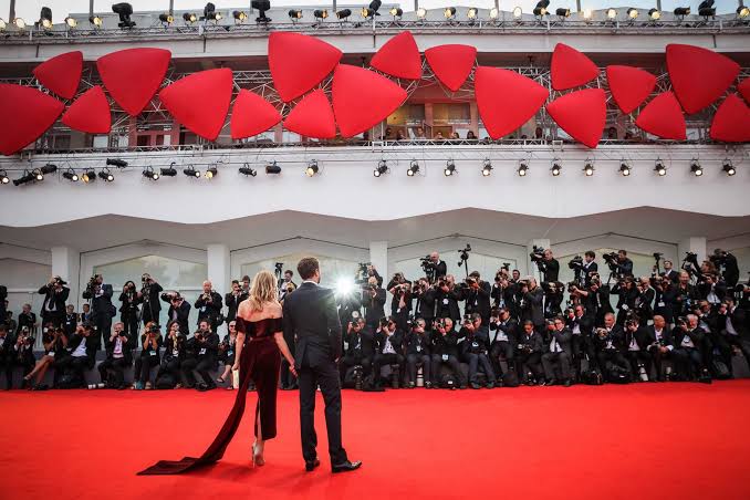 A celebrity couple posing on the red carpet at the Venice International Film Festival, surrounded by a wall of press photographers.