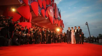 Celebrities and photographers on the red carpet outside the Palazzo del Cinema during the Venice International Film Festival at sunset.