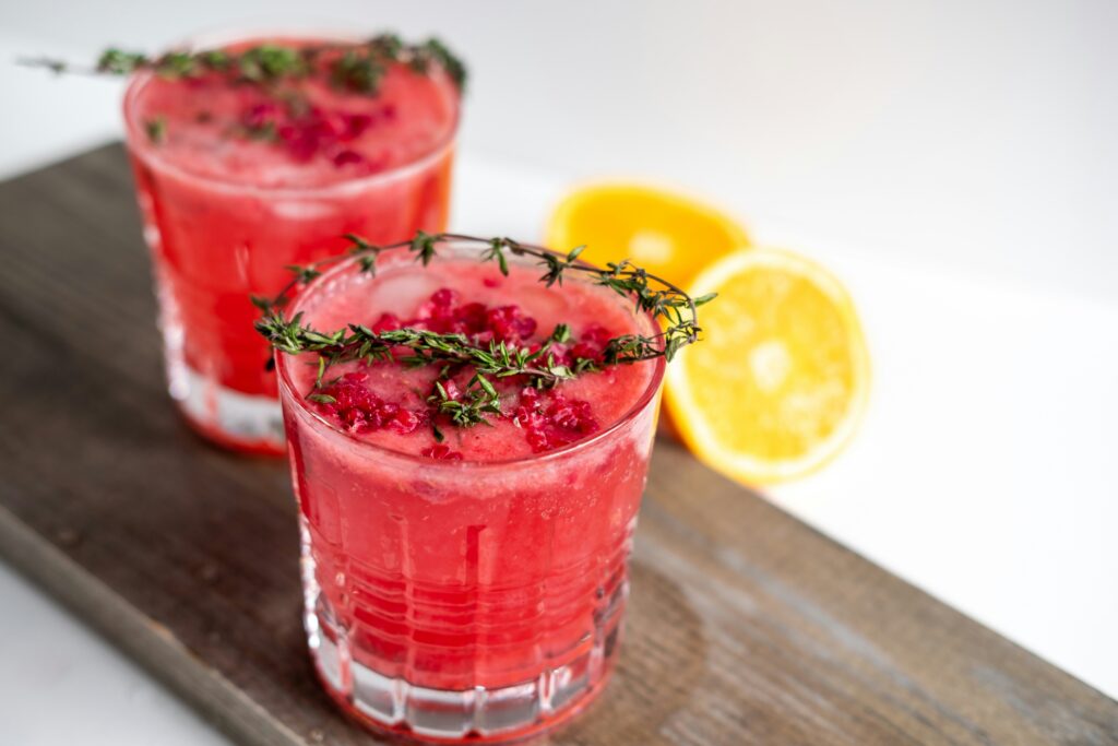 Two pomegranate cocktails garnished with fresh thyme and pomegranate seeds, served on a wooden board