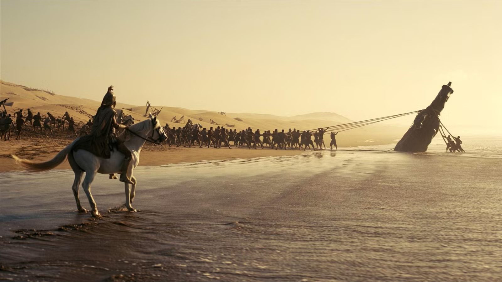 "A Greek warrior on a white horse watching an army pull a massive wooden ship onto a sandy beach, in a cinematic scene from Christopher Nolan's 2026 film, The Odyssey.