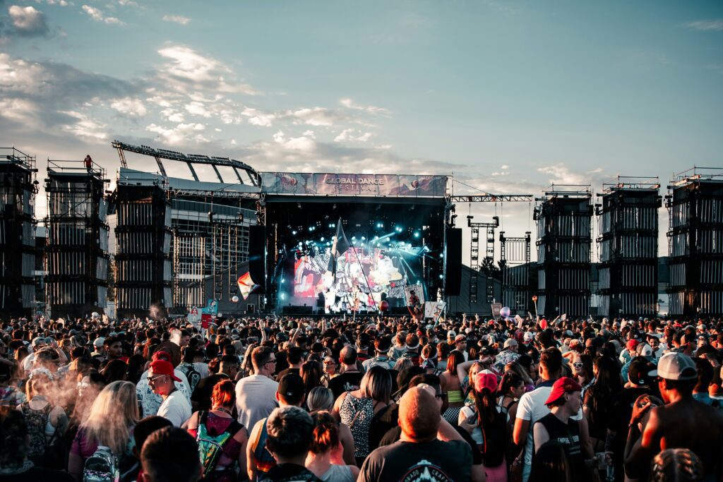 Large crowd at an outdoor electronic music festival in front of a main stage at sunset