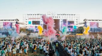 Crowd at an outdoor music festival with colorful powder smoke filling the air in front of a lit stage